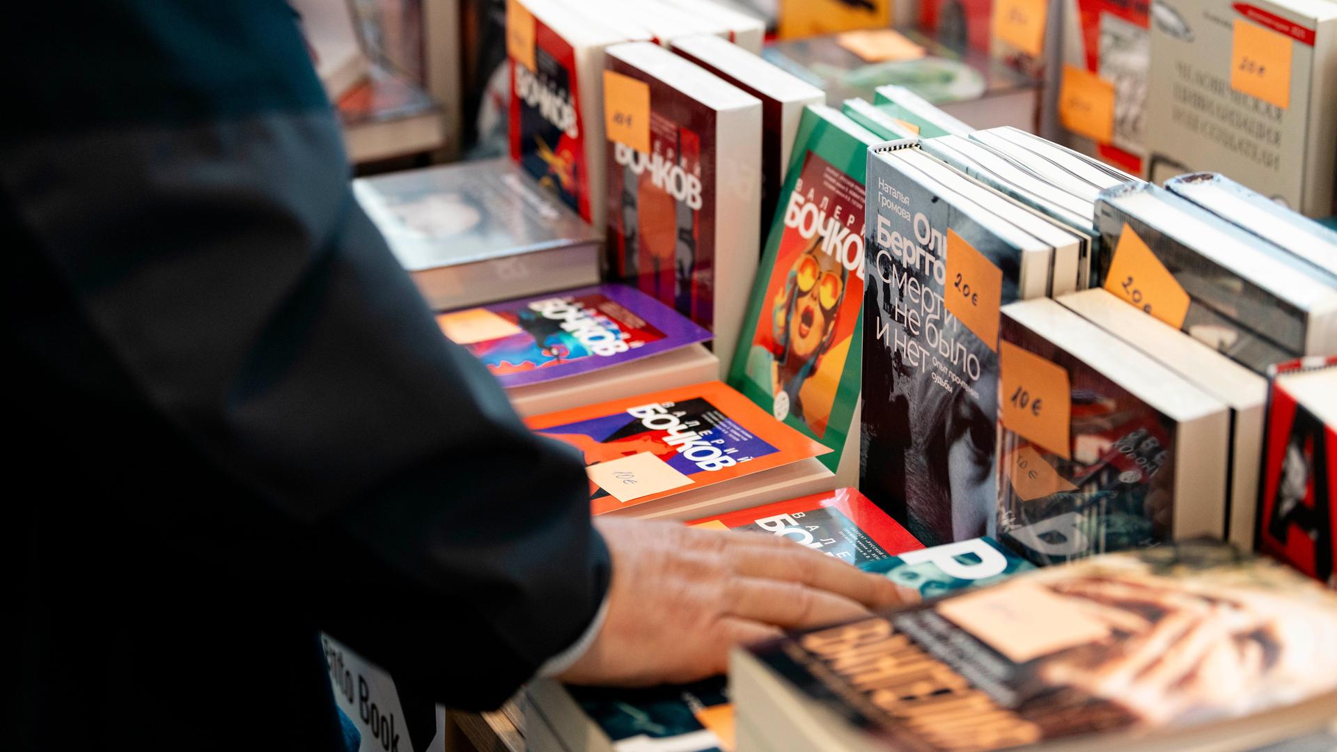 Auf der russischsprachigen Buchmesse in Berlin werden verschiedene russischsprachige Bücher zum Verkauf angeboten. Abgebildet ist ein Tisch, auf dem verschiedene Bücher aufgestellt und gestapelt liegen. Die Hand eines Mannes berührt eines der Bücher.