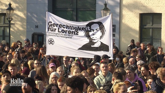 In einer größeren Menschenmenge wird ein großes Banner mit der Aufschrift "Gerechtigkeit für Lorenz" und dem Gesicht des 21-jährigen hochgehalten. © Screenshot 