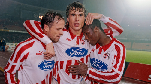 © imago - Das Foto zeigt die drei Fußballspieler Karsten Baumann, Toni Polster und Pablo Thiam (v.l.n.r.) in gestreiften Trikots mit dem Ford-Logo auf der Brust. Es ist das Trikot des 1. FC Köln der Saison 1994-95. Sie stehen eng beieinander und haben ihre Arme umeinander gelegt, als ob sie einen Sieg feiern. Die Spieler tragen weiße Trikots mit roten Streifen und das Puma-Logo ist auf den Ärmeln sichtbar. Im Hintergrund ist ein Stadion zu sehen, das teilweise beleuchtet ist. Es scheint Nacht zu sein oder es könnte sich um ein Spiel unter Flutlicht handeln.