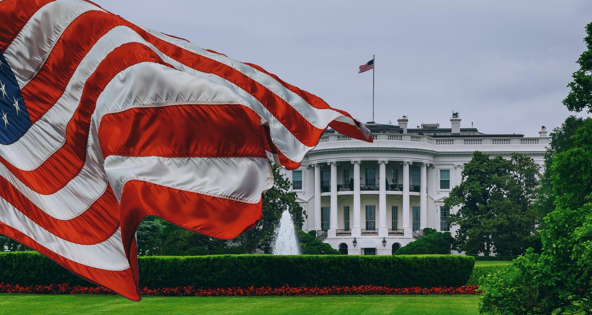 Zu sehen ist das Weiße Haus, im Vordergund weht eine US-Flagge.