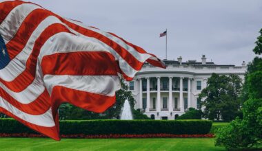 Zu sehen ist das Weiße Haus, im Vordergund weht eine US-Flagge.