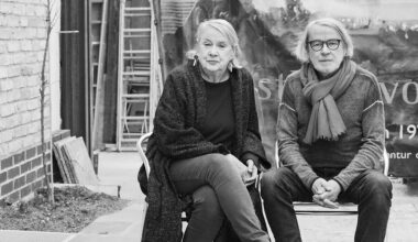 Ute und Werner Mahler sitzen auf zwei Stühlen in einem Innenhof. Sie trägt eine lange schwarze Strickjacke und große Ohrringe. Er trägt einen hellen Schal und eine schwarz umrandete Brille.