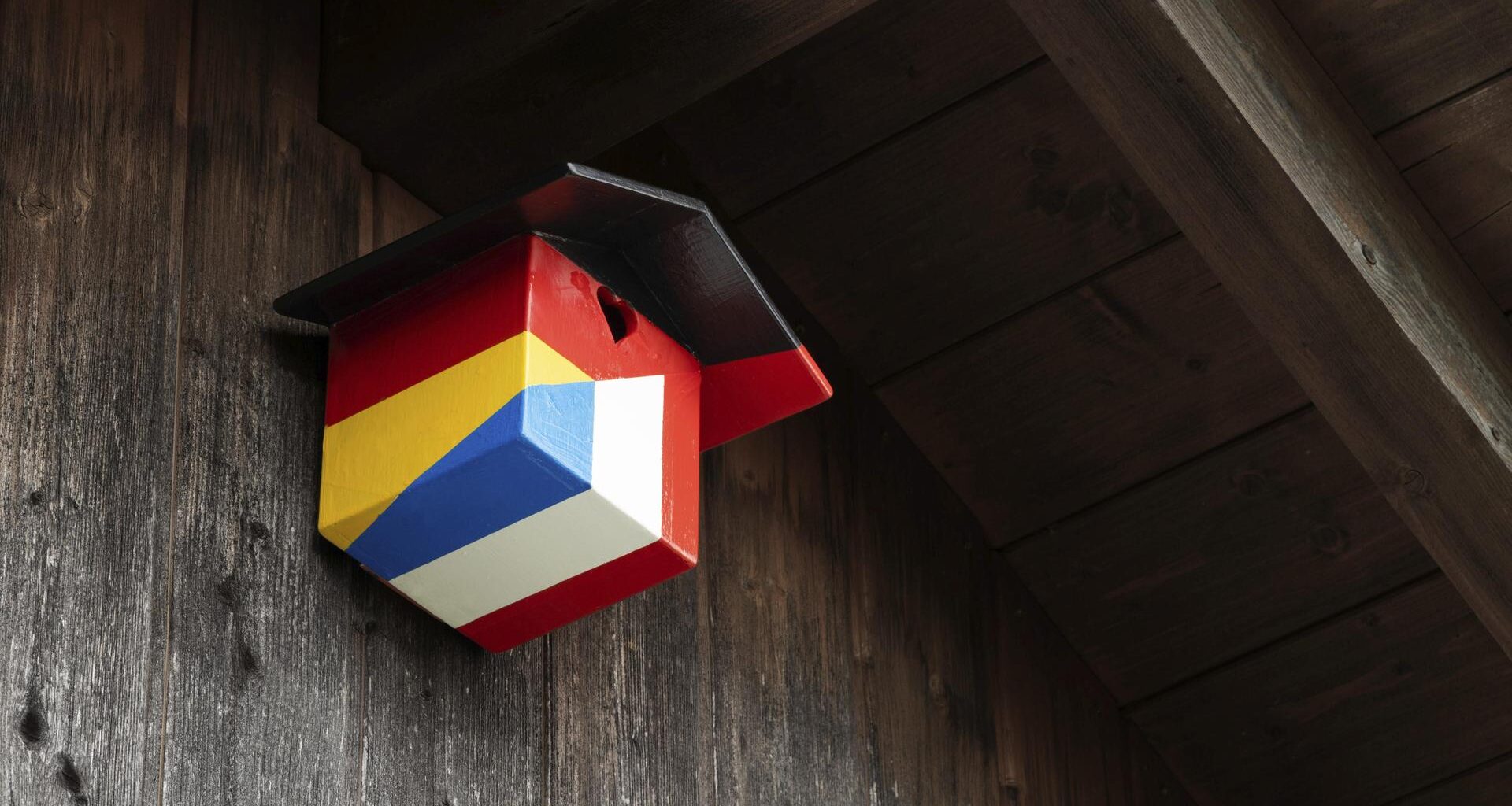 Ein Nistkasten für Vögel in den Farben der deutschen und französischen Nationalflagge hängt an unter einem hölzernen Dachgiebel.
