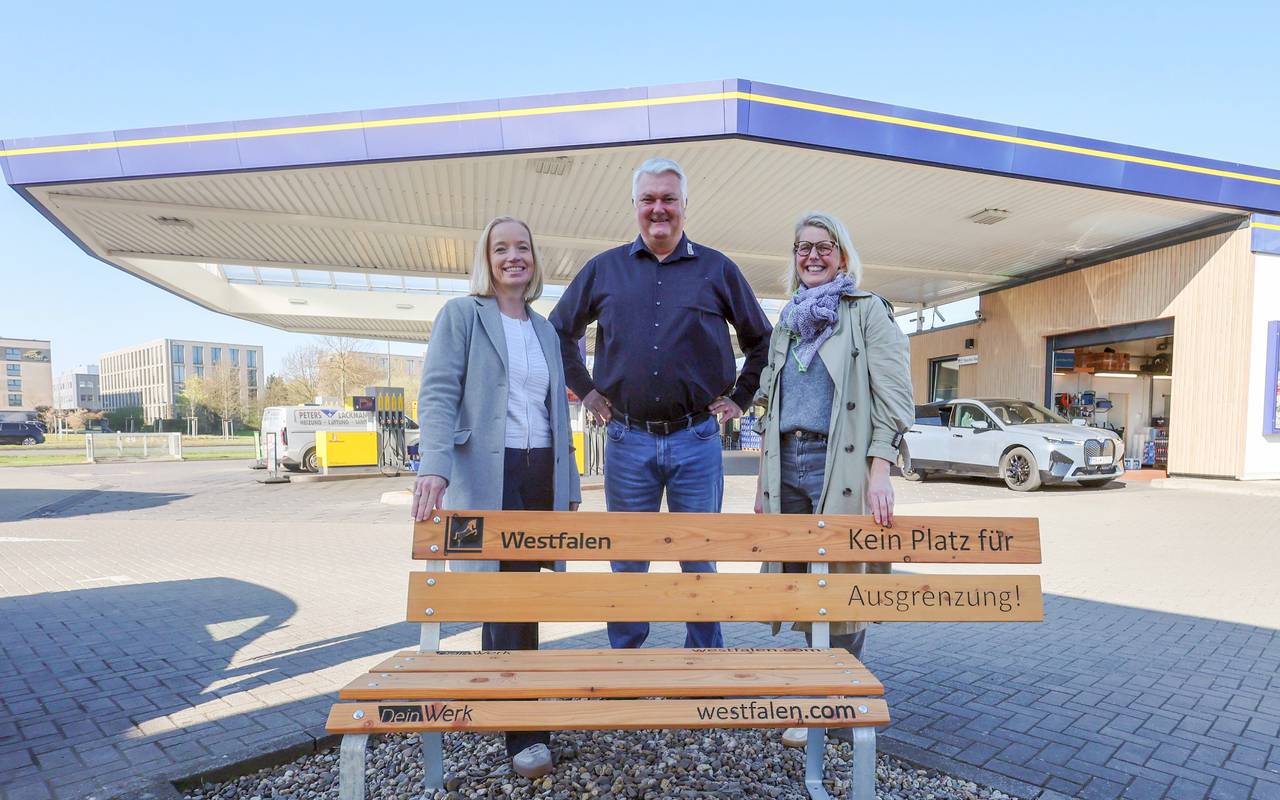 Zwei Frauen und ein Mann hinter einer besonderen Sitzbank an einer Tankstelle.