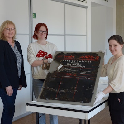 Das Foto zeigt die drei Frauen. Zwei von ihnen halten eine historische Infotafel.