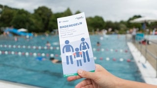Becken im Freibad. Im Vordergrund eine Hand, die einen Flyer mit der Aufschrift "Baderegeln" hält. Darin steht, was erlaubt und was verboten ist.