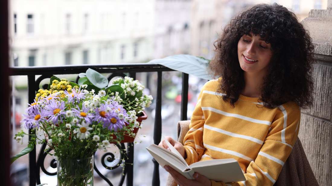 Eine junge Frau liest ein Buch auf einem Balkon (Symbolfoto)