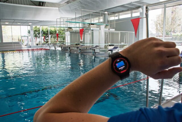 Vor dem Becken eines Hallenbads ist ein Arm mit einer Smartwatch zu sehen, auf der ein Alarm aufleuchtet.