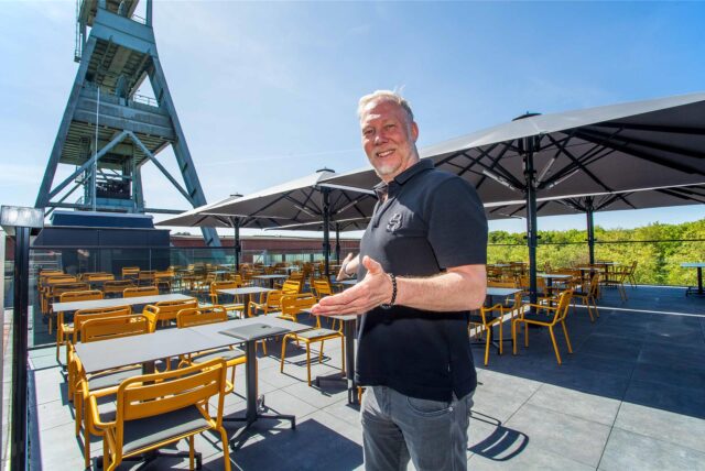 Mitbetreiber Carsten Kubacki steht in der Rooftop-Bar des Restaurants Foodgarage Moto59 in Herten und macht eine einladende Geste mit den Armen.