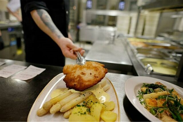 Ein Koch bereitet ein Gericht in einer Restaurantküche zu. Das Gericht besteht aus Schnitzel, Spargel und Kartoffeln, angerichtet auf einem Teller.