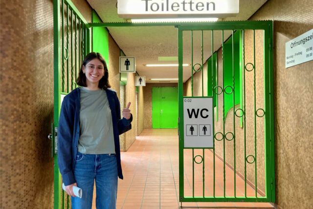 Xenia zeigt Daumen vor dem Eingang zur Toilettenanlage Kampstraße Daumen hoch. Denn die Anlage ist sehr sauber.