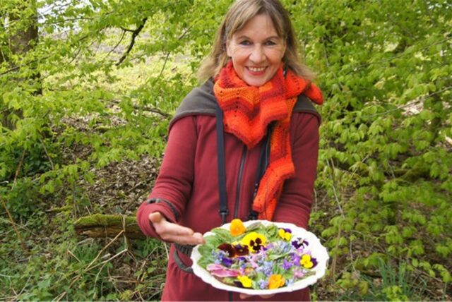 Ursula Stratmann steht im Wald und hält einen Teller mit Blüten und Kräutern in die Kamera.