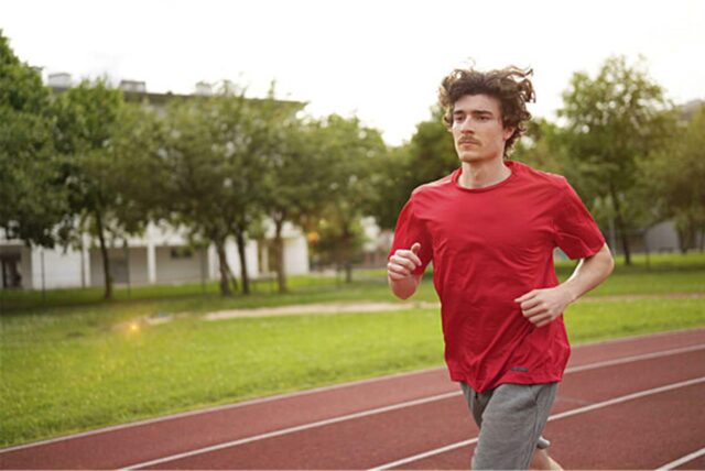 Ein junger Mann in Sportkleidung joggt auf einer roten Laufbahn. Im Hintergrund sind Bäume und ein Gebäude zu sehen.