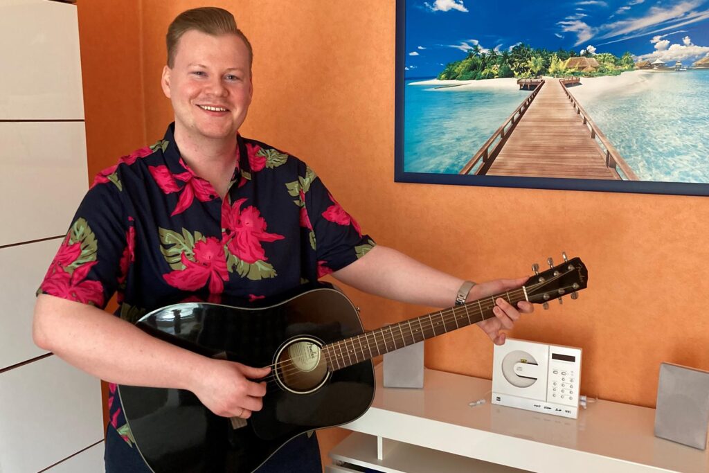 Der Hertener Schlagersänder Steven Loyd steht in einem Wohnzimmer und hält eine Gitarre in den Händen.