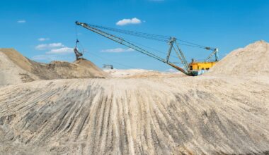 Ein großer industrielle Bagger-Maschine Sand auf einem Hügel in einem Tagebau. Im Vordergrund befindet sich rötliches Wasserteich.