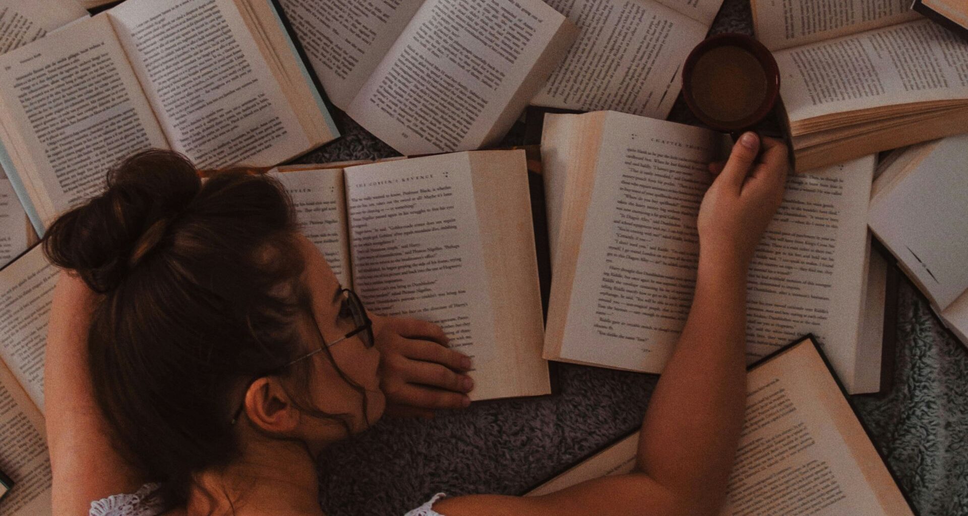 Frau liegt auf vielen offenen Bücher und hat einen Kaffe in der Hand.