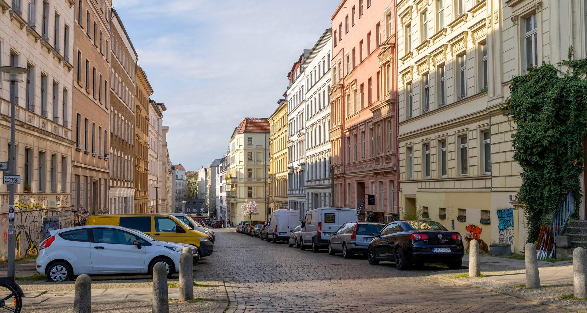 Eine Straße mit schönen Altbauwohnungen in Berlin. Auf der Straße parken ein paar Autos. Vor einem Haus im Vordergrund ist auch ein kleiner, grüner Baum oder Strauch.