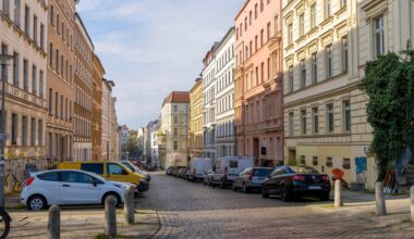 Eine Straße mit schönen Altbauwohnungen in Berlin. Auf der Straße parken ein paar Autos. Vor einem Haus im Vordergrund ist auch ein kleiner, grüner Baum oder Strauch.