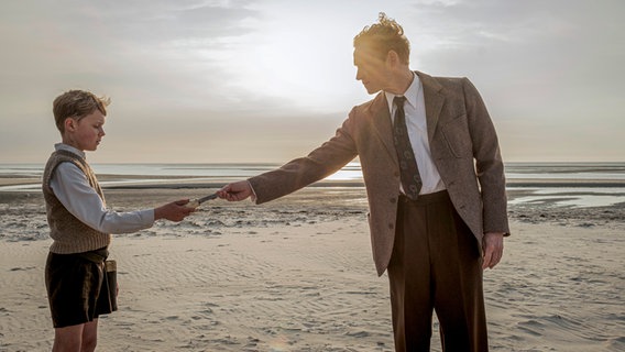 Ein Junge steht am Strand und reicht einem Mann etwas in die Hand  - Filmszene mit Matthias Schweighöfer (r.) und Jasper Billerbeck aus dem Film "Amrun" © 2025 bombero international GmbH & Co. KG / Rialto Film GmbH / Warner Bros. Entertainment GmbH / Mathias Bothor Foto: Mathias Bothor