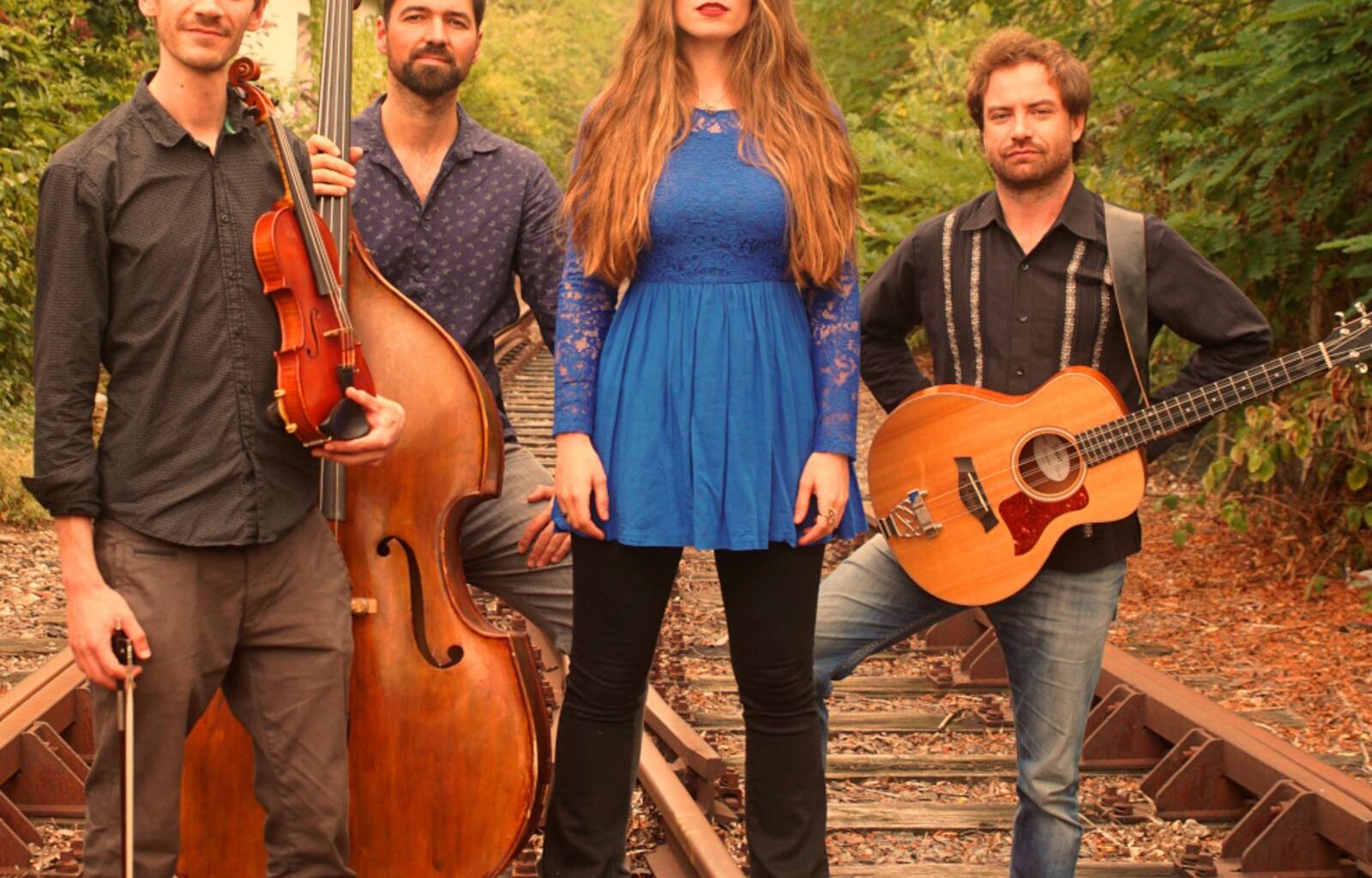 Eine Band, bestehend aus drei Musikern und einer Musikerin, steht in einem Gleisbett. Dabei sind die Instrumente Kontrabass, Gitarre und Violine.