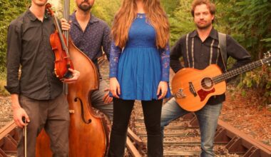 Eine Band, bestehend aus drei Musikern und einer Musikerin, steht in einem Gleisbett. Dabei sind die Instrumente Kontrabass, Gitarre und Violine.