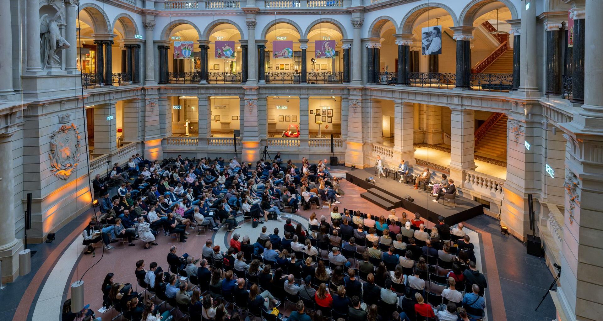 Blick von oben auf das 400köpfige Publikum und eine Bühne im Lichtsaal des Berliner Museums für Kommunikation, umrandet von Galerien und Säulen