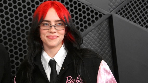 Eine junge Frau mit langen glatten Haaren (oben knallrot, unten schwarz) lächelt vor einer Wand (Musikerin Billie Eilish) © IMAGO / UPI Photo 