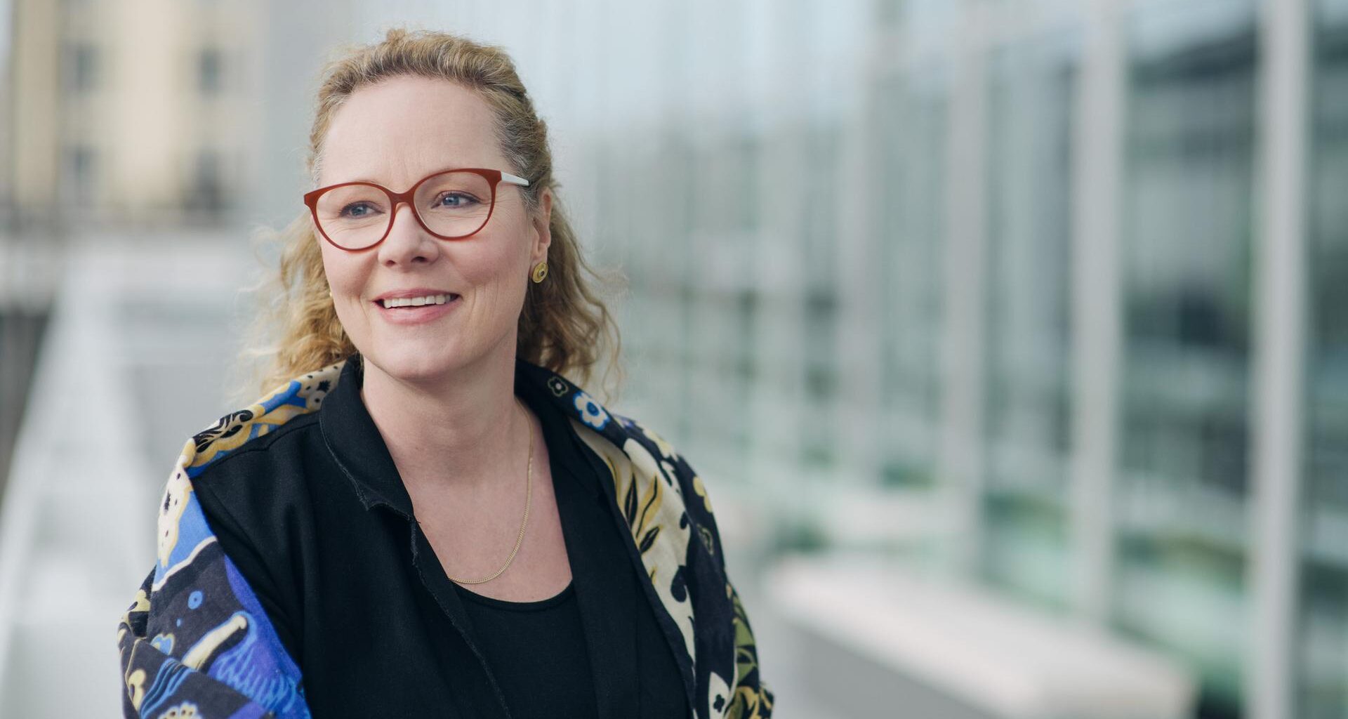 Eine Frau mit blondem Haar steht vor einer Glasfassade. Sie trägt eine dunkelrote Brille und blickt lächelnd links aus dem Bild heraus. Sie trägt ein schwarzes Oberteil und eine geblümte Jacke darüber.