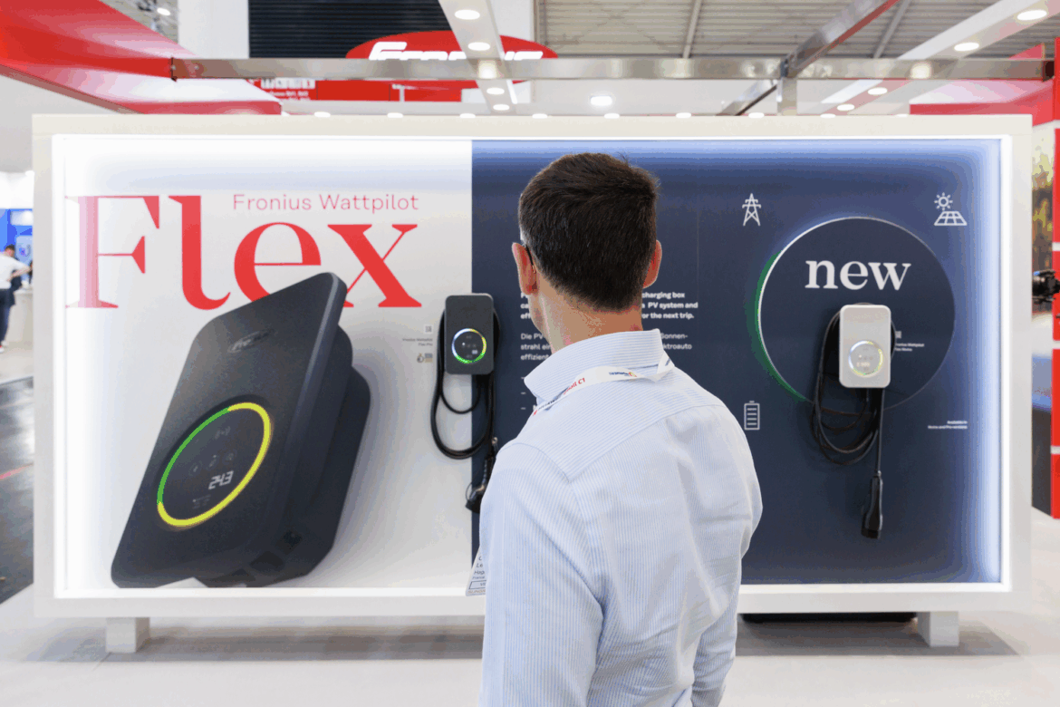 Ein Mann steht vor einer Messewand mit zwei E-Ladegeräten des Typs Fronius Wattpilot Flex. Links ist das Produkt groß abgebildet, rechts ein neues Modell montiert.