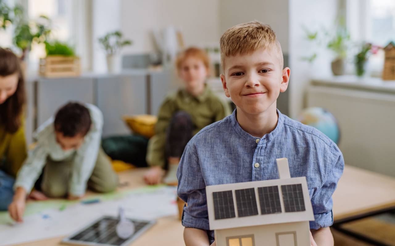 Ein Junge hält ein gebasteltes Holzhaus mit Solaranlagen auf dem Dach in die Kamera und lächelt. Im Hintergrund weitere Schulkinder, die an einem Tisch an etwas arbeiten.