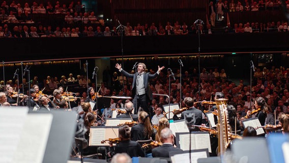 Der Dirigent Stanislav Kochanovsky steht vor einem Orchester, im Hintergrund ein Saal voller Publikum. © NDR Foto: Michael Neugebauer