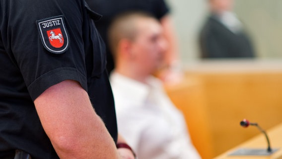 Ein Angeklagter im Gerichtssaal; im Vordergrund ist die Aufschrift "Justiz" auf dem Hemd eines Beamten zu sehen. © dpa Foto: Julian Stratenschulte