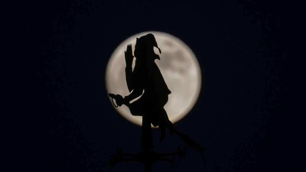 Der fast vollständige Mond steht hinter einer Wetterfahne in Form von einer auf einem Besen reitenden Hexe.