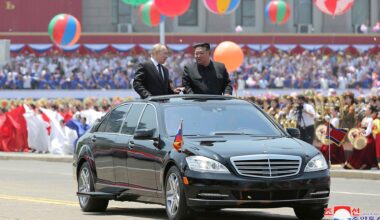 Russlands Präsident Putin und Nordkoreas Machthaber Kim Jong Un stehen im geöffneten Dachfenster eines schwarzen Autos, das durch eine Willkommensparade in Pjöngjang fährt. Im Hintergrund sind bunte Ballons und viele Menschen zu sehen.