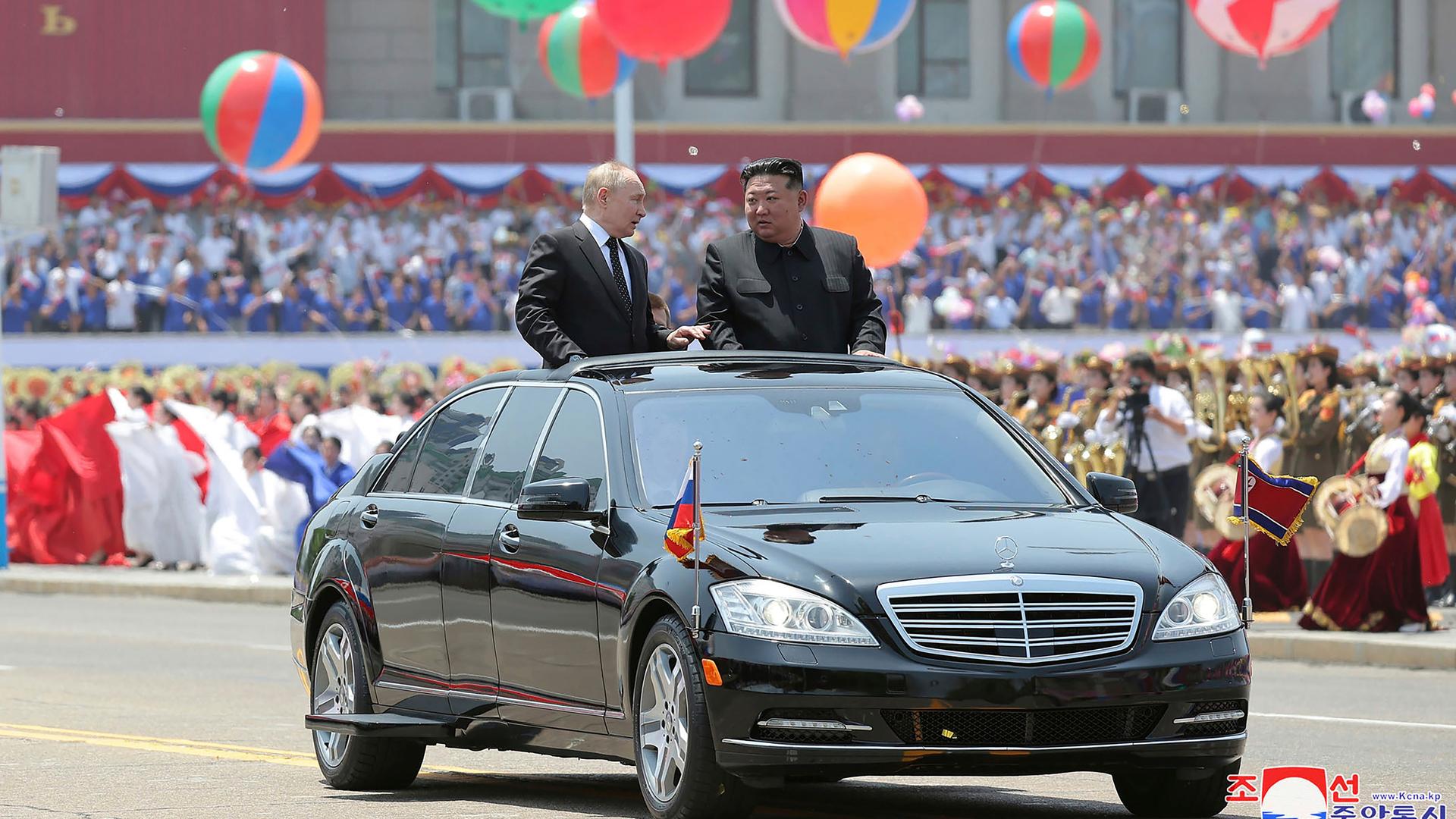 Russlands Präsident Putin und Nordkoreas Machthaber Kim Jong Un stehen im geöffneten Dachfenster eines schwarzen Autos, das durch eine Willkommensparade in Pjöngjang fährt. Im Hintergrund sind bunte Ballons und viele Menschen 