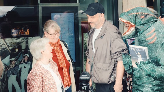 Zwei ältere Frauen lächeln einem Mann mit drei Tage Bart und Baseballmütze zu. Rechte hinten im Bild steht ein grüner Dino udn hält das Opern Programmheft für die neue Spielzeit in der Hand. © Staatsoper Hamburg Foto: Manuel Braun