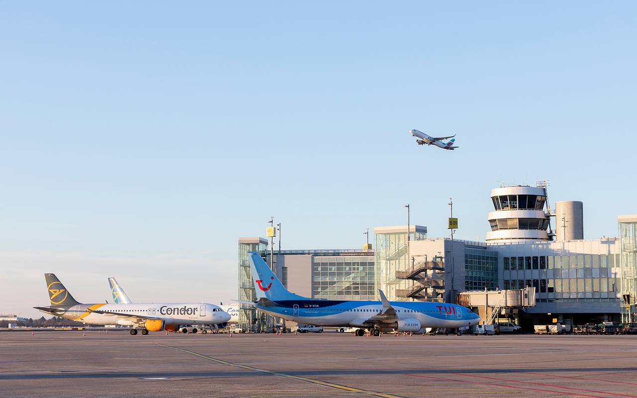 Eine Aufnahme vom Rollfeld des Düsseldorfer Flughafens. Zwei Flugzeuge stehen geparkt vor dem Flughafengebäude. Ein Flugzeug befindet sich in der Luft und startet gerade. 