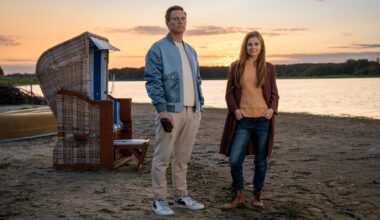 Ein Mann und eine Frau stehen in der Abenddämmerung an einem Strand. Neben ihnen ein Strandkorb, hinter dem Wasser liegt Wald. Er hat eine blaue Jacke über einem weißen Shirt, sie trägt eine rote Jacke über einem orangefarbenen Pullover.