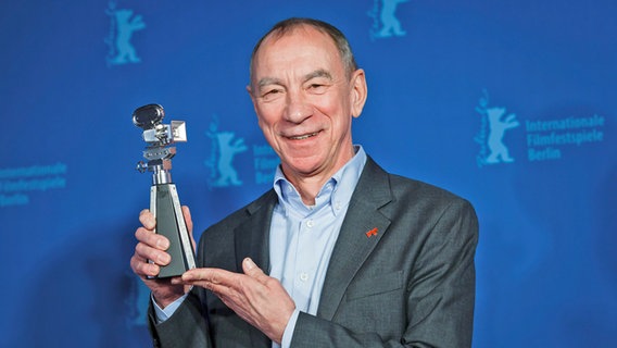 Ein lächelnder Mann hält eine Auszeichnung hoch vor einer blauen Wand der Berlinale - Dr. Rainer Rother hält lächelnd die Berlinale-Kamera in die Höhe © Dirk Deckbar Foto: Dirk Deckbar