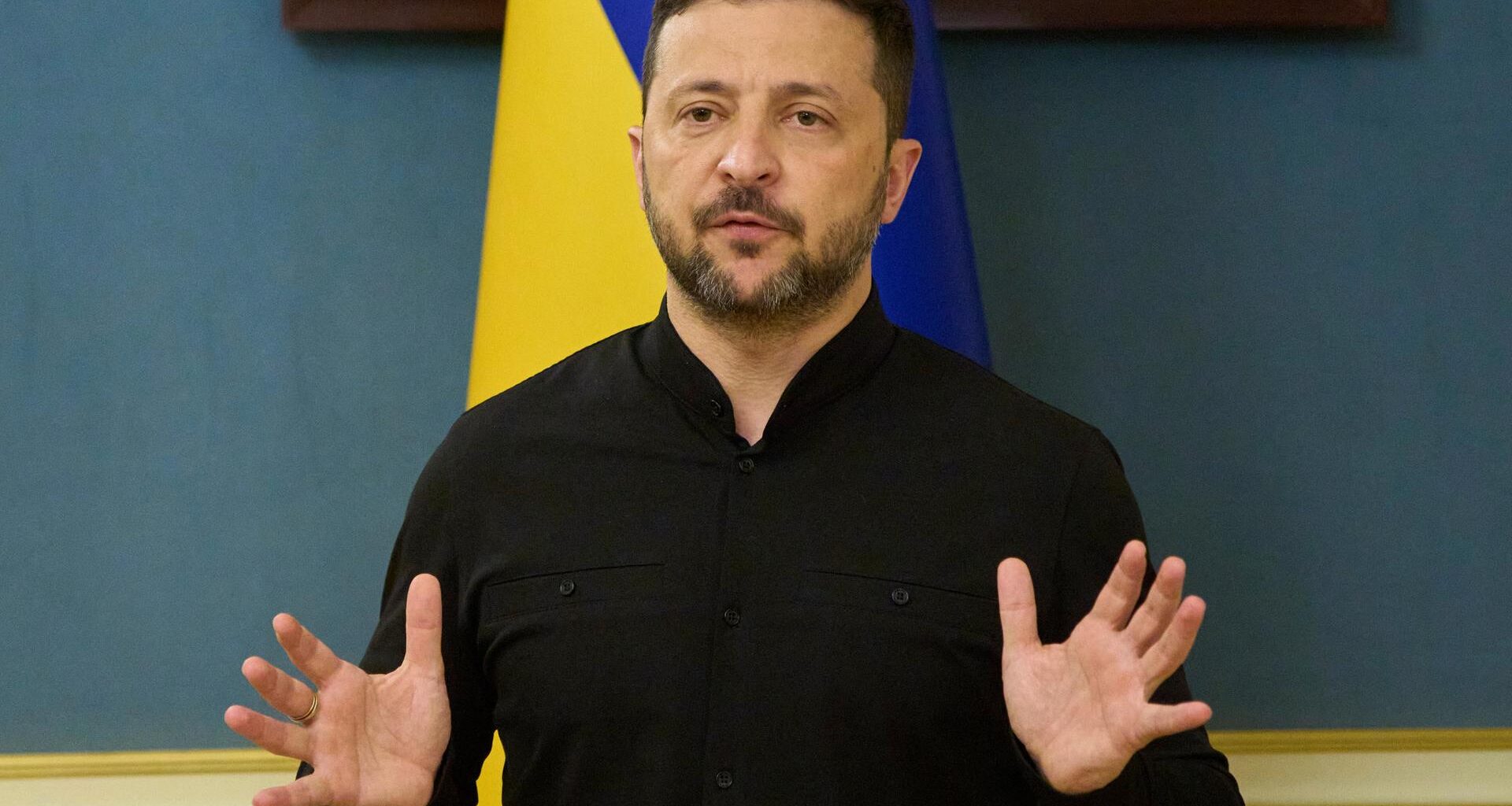 Der ukrainische Präsident Wolodymyr Selenskyj steht vor einer Flagge seines Landes und gestikuliert mit den Händen.