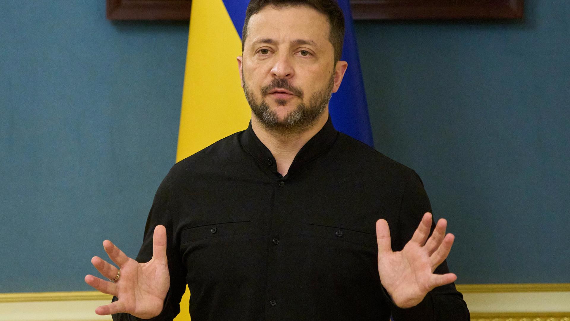 Wolodymyr Selenskyj steht vor einer blau-gelben ukrainischen Landesflagge und hält eine Ansprache, während er mit beiden Händen gestikuliert.