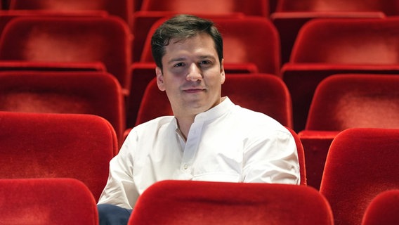 Ein Mann mit weißem Hemd (Demis Volpi) sitzt in einem Theatersaal mit roten Stühlen. © picture alliance / dpa Foto: Marcus Brandt