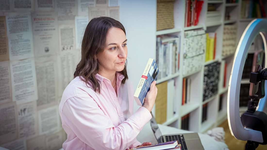 Helen Daughtrey bespricht von ihrer Wohnung in Bonn aus in ihrem virtuellen Buch-Club Bücher. Zu sehen ist Daughtrey vor einer Kamera, sie hält Bücher in den Händen, im Hintergrund ein Bücherregal.