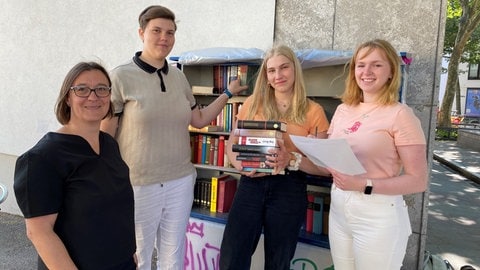 Vier Frauen stehen vor dem Bücherschrank und Mainz-Mombach. Sie halten Bücher und Zettel in der Hand.