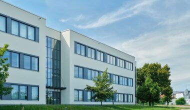 Ein modernes Bürogebäude mit großen Fenstern und einer Glasfassade, umgeben von Bäumen und einem blauen Himmel im Hintergrund.