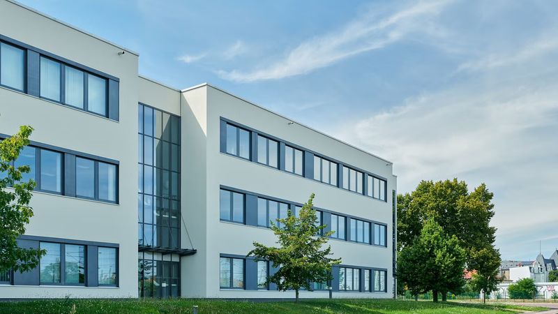 Ein modernes Bürogebäude mit großen Fenstern und einer Glasfassade, umgeben von Bäumen und einem blauen Himmel im Hintergrund.