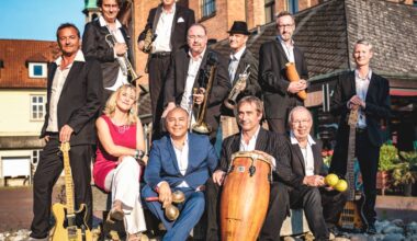 11 Personen stehen oder sitzen mit Instrumenten unter freiem Himmel vor einem historischen Gebäude.