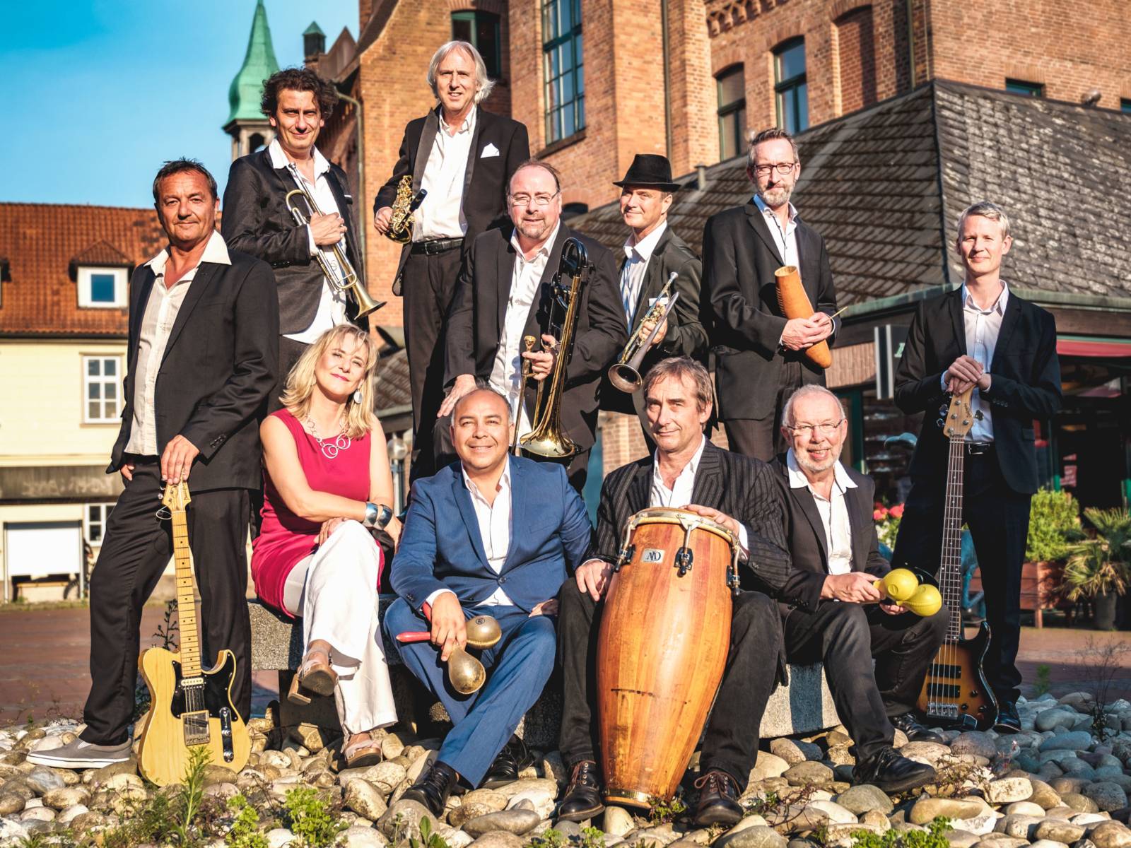11 Personen stehen oder sitzen mit Instrumenten unter freiem Himmel vor einem historischen Gebäude.
