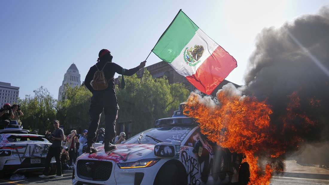 Alles begann mit einer Razzia der US-Einwanderungsbehörde ICE, jetzt brennen Autos und die Nationalgarde ist im Einsatz. In Los Angeles demonstrieren seit Samstag Menschen überwiegend friedlich, teils gewaltsam, gegen die das ICE. Im Foto steht ein Demonstrant auf einem brennenden Waymo-Taxi in der Nähe des Metropolitan Detention Center in der Innenstadt von Los Angeles. 
