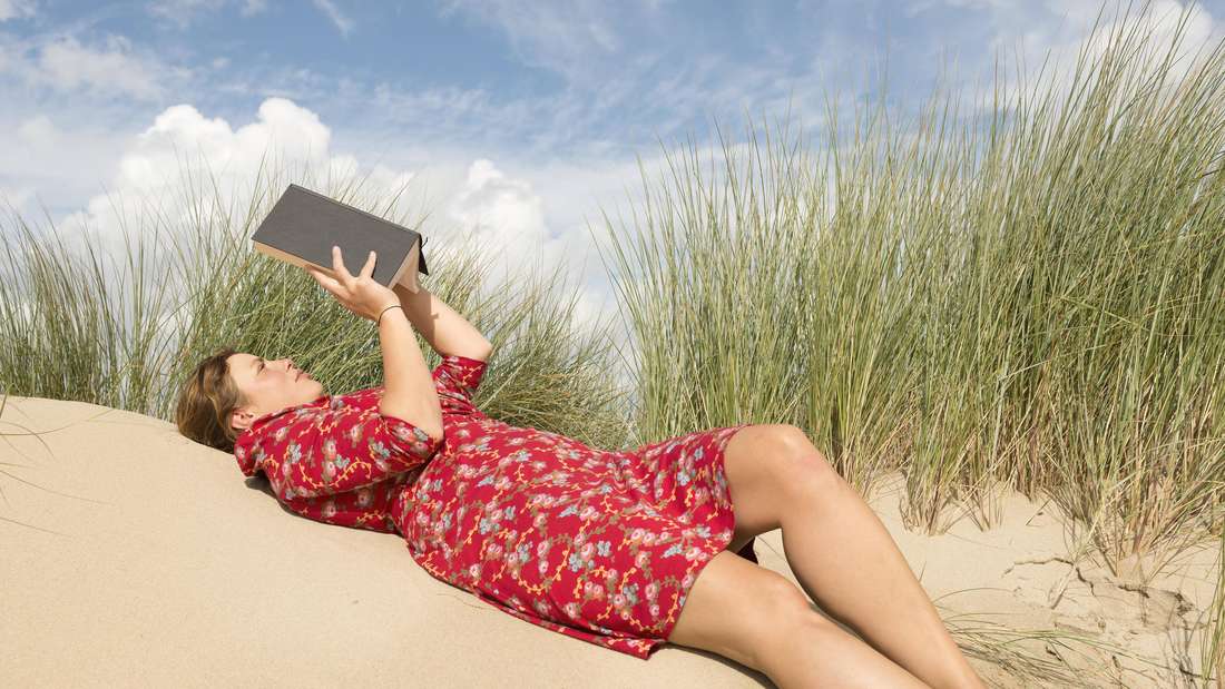 Eine Frau liest ein Buch in einer Düne am Strand (Symbolfoto)